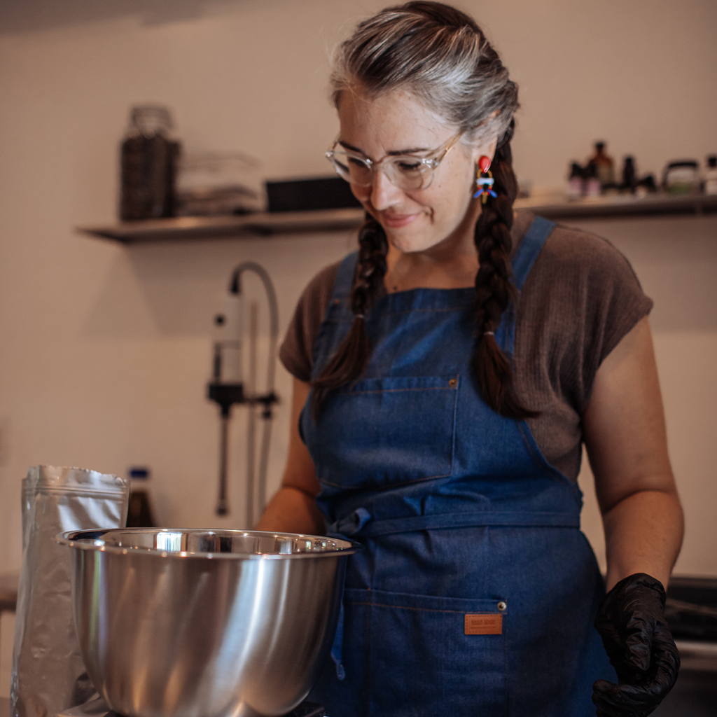 Little Bird's formulator, Dyana, is standing in front of a scale measuring ingredients in her studio lab. There is a shelf behind her with jars and equipment. She is wearing a denim apron and her hair is braided.