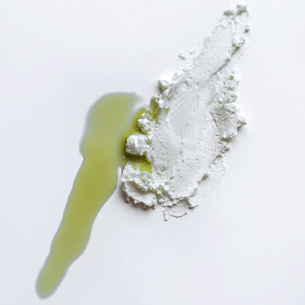On a white surface there is a smear of green oil next to a smear of white cream to show texture and color. The cream looks thick and opaque and the oils is runny and bright green.